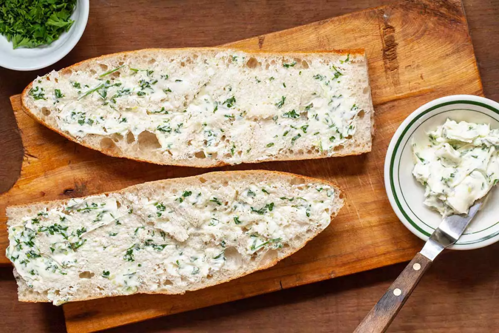 Cắt miếng bánh mì làm đôi theo chiều dọc, sau đó lấy một cái chén bỏ bơ, tỏi, và rau mùi tây vào cùng trộn đều ta được hỗn hợp sau đó bỏ trét lên toàn bộ bề mặt bánh.