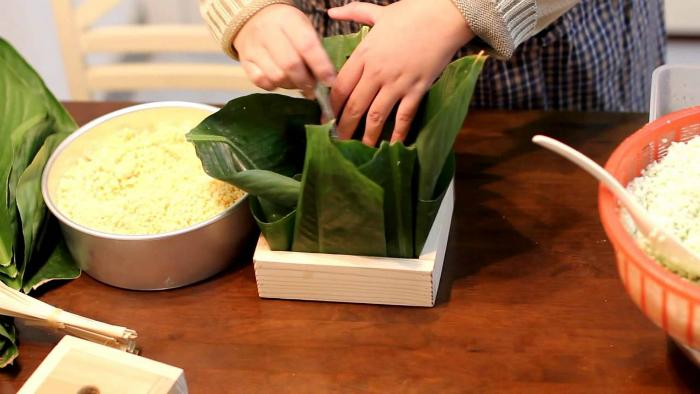 Cách gói bánh chưng vuông bằng khuôn
Bạn hãy chuẩn bị một chiếc khuôn hình vuông để bánh được đều và đẹp hơn.
Đầu tiên bạn xếp 4 lá dong bằng cách gấp mép dưới lên, gấp mép bên trái qua để tạo đường nếp cho lá sau đó đặt 4 lá đã được gấp xuống dưới khuôn rồi đổ gạo nếp lên trên