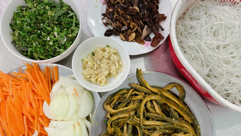 Nguyên Liệu Làm Miến Xào Lươn
Các bạn hãy chuẩn bị theo danh sách nguyên liệu để làm miến xào lươn dưới đây:
Lươn đồng: 500g (nên chọn lươn đồng tươi, da vàng óng, thịt săn chắc)
Miến dong: 200g (chọn miến sợi nhỏ, suôn thẳng, màu hơi đục xám)
Giá đỗ: 200g
Nấm hương: 4-5 cánh
Mộc nhĩ: 1 tai
Hành tây: 1 củ
Ớt sừng: 1 quả
Rau răm: 1 bó
Hành lá: 1 nắm
Trứng gà: 1 quả
Nước hầm xương: 1 bát tô (tùy chọn)
Gia vị: Mắm, muối, hạt tiêu, hành khô
Mỡ lợn hoặc dầu ăn
Bột chiên giòn
Hành phi: 1 ít